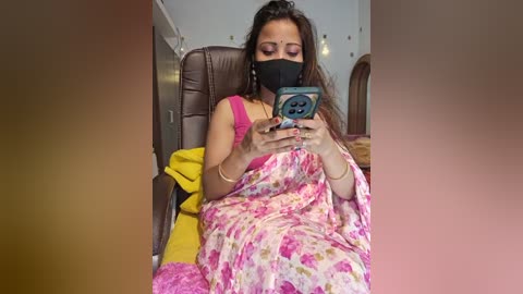 Media: Video of a woman in a pink floral sari with a black face mask, holding a phone, sitting in a brown leather chair, with yellow and white background.