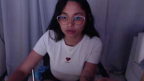 Media: Video of an Asian woman with long black hair, glasses, and a white T-shirt with a heart design. She's seated indoors with white curtains in the background, holding a phone.