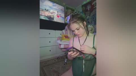 Media: A young, pale-skinned woman with blonde hair, wearing a green lingerie set, sits on a bed, absorbed in a smartphone. The room features pastel-colored walls, a pink chair, and a TV displaying a beach scene.