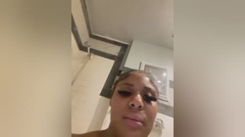 Media: A video of a young Black woman with dark skin, braided hair, and wearing a light-colored top, standing in a beige-tiled bathroom with a framed picture on the wall.