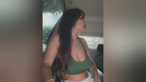 Video of a light-skinned woman with long, dark hair, wearing a green sports bra and white shorts, sitting in a car with a green tree visible outside.