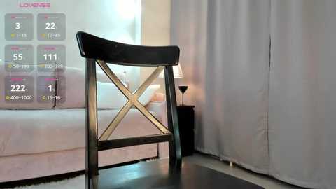 Video of a minimalist bedroom with a black wooden stool with crossbar backrest, white curtains, and a white bed in the background.