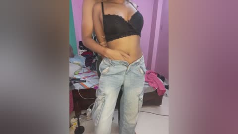 Media: Video of a slender, light-skinned woman with medium-length hair, wearing a black lace bra and ripped light blue jeans, standing in a cluttered bedroom with colorful clothing on the bed.