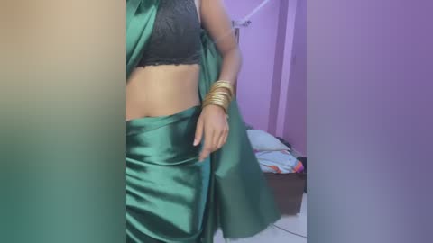 Media: Video of a woman with medium brown skin, wearing a black lace bra, green satin sari, and gold bangles, standing in a room with purple walls and a bed with colorful sheets.