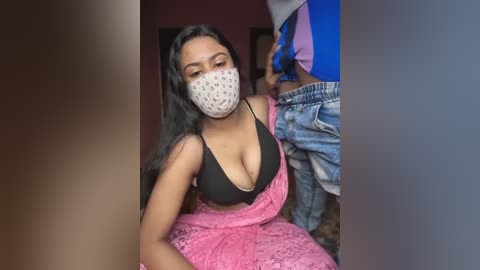 Media: Video of a woman with long black hair, wearing a black bra, pink lace dress, and face mask, sitting on a bed. A man in blue jeans and a blue shirt is partially visible.