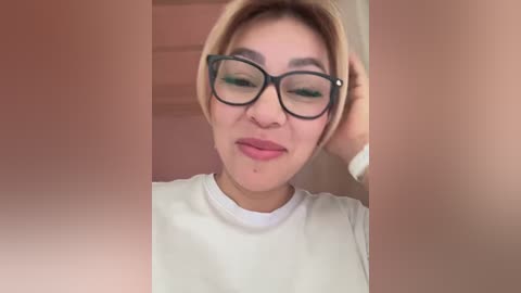 Media: Video of an Asian woman with short, light brown hair, wearing black-framed glasses, a white t-shirt, and a watch, with a pimple on her cheek.