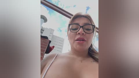 Media: Video of a plus-size, light-skinned woman with glasses, wearing a white bra, standing on a balcony with a cloudy sky and city buildings in the background.