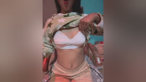 Media: Video of a light-skinned woman with medium-length dark hair, lifting a patterned shirt to reveal a white bra. She wears plaid pajama pants. Background shows a bed with a patterned blanket and a red object.