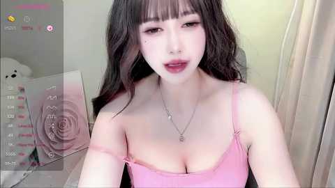 Media: Video of an East Asian woman with long black hair, wearing a pink spaghetti-strap top, standing indoors. She has fair skin and is smiling, with makeup including pink lipstick. Background includes a white wall and a teddy bear.