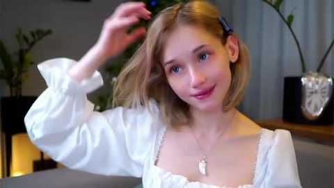 Media: Video of a young woman with light skin and shoulder-length blonde hair, wearing a white blouse with puffed sleeves, posing indoors with a potted plant and decorative objects in the background.