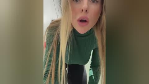 Media: Video of a blonde woman with fair skin, wearing a green turtleneck sweater, leaning forward with a surprised expression, set against a blurred beige background.