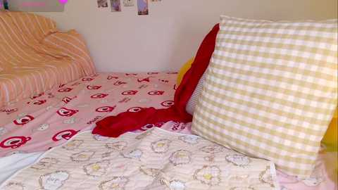 Video of a cozy, cluttered bedroom with a pink bedspread adorned with red and white circular patterns. A red blanket and two pillows with yellow and white checkered covers lie on the bed.