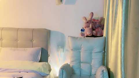 Media: Video of a cozy, pastel-colored bedroom featuring a plush, light blue armchair with teddy bears and a soft, tufted beige headboard. The room is illuminated by a soft, warm light.