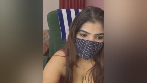 Media: Video of a woman with long brown hair, wearing a blue patterned face mask, green chair, and striped towel, blurred background.