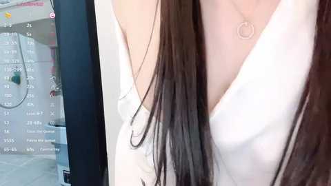 Media: Video of a woman with long, straight brown hair, wearing a white, off-the-shoulder dress. She stands in a modern bathroom with white tiles, a shower, and a door visible in the background.