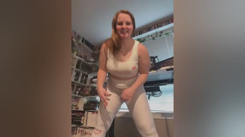 Media: Video of a smiling, fair-skinned, blonde woman wearing white crop top and pants, bending over in a cluttered, book-filled room.