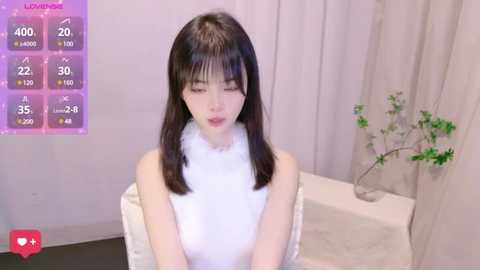 Media: Video of a young, pale-skinned, slim Asian woman with shoulder-length straight black hair, wearing a sleeveless white dress, sitting on a white chair in a minimalist room with white walls and a vase of green plants.