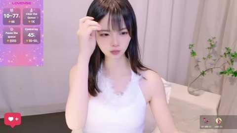 Media: Video of a young East Asian woman with straight black hair, wearing a white lace sleeveless top, looking worried. Background features a white couch, a vase with greenery, and a digital thermometer overlay.