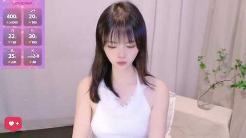 Media: A video of a young, fair-skinned Asian woman with long, straight black hair, wearing a white tank top, sitting indoors with a white table and green plant in the background.