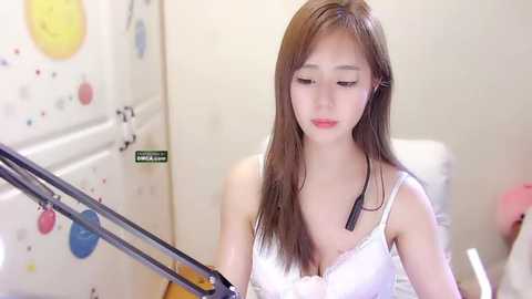 Media: A video of a young Asian woman with straight, long brown hair, wearing a white lace bra, sitting at a piano in a brightly lit room with colorful wall decorations.