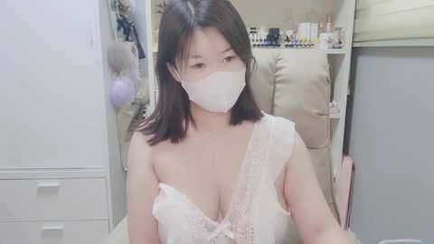 Media: Video of an East Asian woman with medium skin tone and straight, shoulder-length dark hair, wearing a white lace bralette and face mask, seated in a beige recliner in a clean, white room with shelves of toiletries in the background.