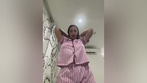 Video of a woman with medium brown skin, wearing pink and white striped pajamas, standing in a modern room with white walls and a black and white floral wallpaper.