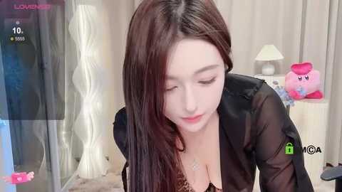 Media: A video of a young Asian woman with long dark hair, wearing a sheer black top, bending over. Background includes a lamp, pink Kirby plush toy, and curtains.