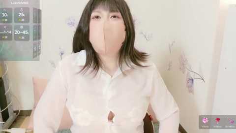 Media: A video of an East Asian woman with straight black hair, wearing a sheer white blouse, partially covering her face with a pink face mask. The background shows a minimalist room with white walls and floral decals.