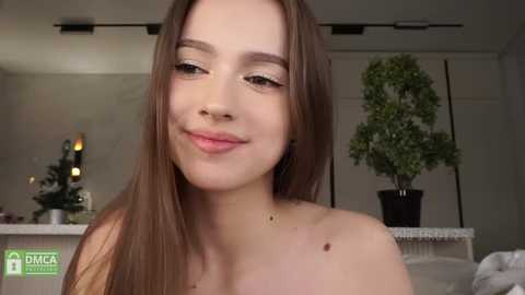 Video of a young, light-skinned woman with long brown hair, smiling, in a softly lit room with a Christmas tree, potted plant, and a white wall.