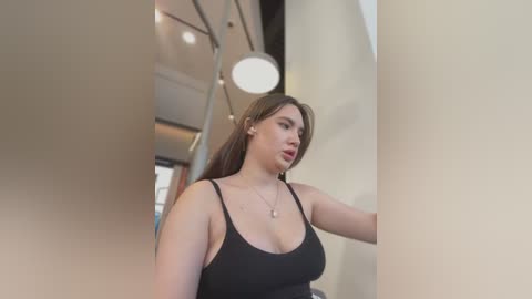 Media: Video of a young woman with long brown hair and light skin, wearing a black tank top, standing in a modern, well-lit room with beige walls and a spherical light fixture.