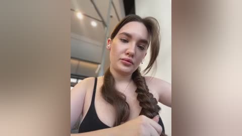 Media: Video of a young woman with fair skin, brown hair in a braid, wearing a black tank top, indoors with modern, minimalist decor.