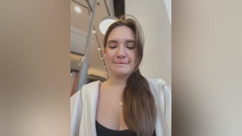 Media: Video of a smiling young woman with long brown hair, wearing a black top and a white cardigan, standing indoors near a modern, beige wall and lamp.