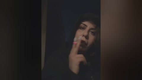 Media: A dimly lit video of a person with a dark complexion, wearing a black hat, smoking a cigarette, and covering their mouth with their index finger, standing in a dark, shadowy doorway.