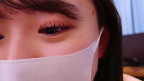 Media: Video of an Asian woman's face, showing her light skin, dark brown eyes, and a white surgical mask partially covering her nose and mouth. The background is blurred, suggesting an indoor setting.