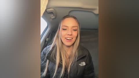 Media: A video of a smiling, fair-skinned, blonde woman with long hair, wearing a black North Face jacket, seated in the backseat of a car. The background shows a light-colored interior with a beige headrest.