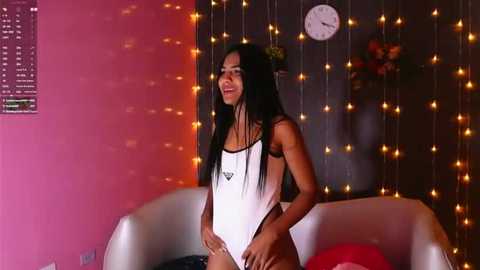 Media: Video of a slim, young woman with long black hair, wearing a white one-piece swimsuit, standing confidently in a dimly lit room with warm fairy lights, a clock, and a modern sofa.