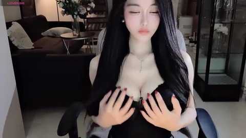 Media: Video of an Asian woman with long black hair, wearing a black dress, covering her breasts with her hands in a dimly lit living room.