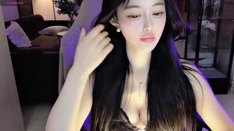 Media: A video of an East Asian woman with long, straight black hair, wearing a black lace bralette and adjusting her hair in a modern, dimly-lit living room with a sofa and plants in the background.