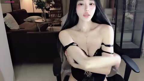 Media: Video of a slender, East Asian woman with long black hair, wearing black lingerie, sitting in a modern living room with a dark sofa and bookshelves.