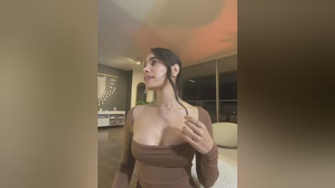 Video of a young woman with medium skin tone, dark hair in a ponytail, wearing a strapless brown top, standing indoors with a blurred background featuring a modern, dimly lit room with wooden floors and large windows.