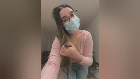 Media: Video of a young woman with long brown hair, wearing glasses, a light blue face mask, and a pink cardigan, taking a selfie indoors.