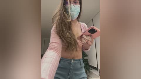 Media: Video of a young woman with long brown hair, wearing a pink cardigan, blue jeans, and a face mask, taking a selfie in a dimly lit room.