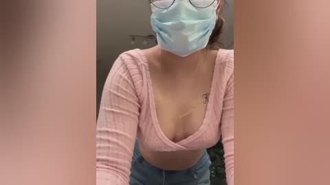 Media: Video of a young woman in a light pink ribbed crop top, revealing a small tattoo on her chest, wearing a blue face mask and glasses. She is in a dimly lit room, leaning forward.