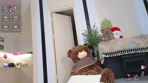 Media: Video of a cozy room with a brown teddy bear, green Christmas tree, and Santa Claus plush toy, all on a white chair, against a white wall with a door.