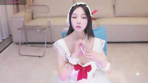 Media: Video of an Asian woman in a white maid outfit with a red bow, kneeling on a white floor, holding a pink feather duster, with a beige couch and chair in the background.