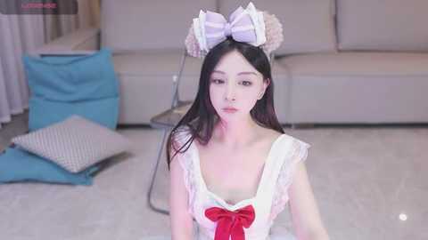 Media: A video of a young Asian woman with long black hair and fair skin, dressed in a frilly white maid costume with a large pink bow, sitting on a light-colored carpeted floor.