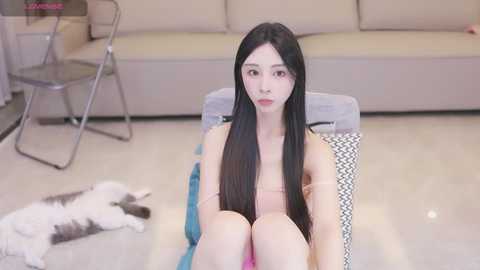 Media: Video of a young East Asian woman with long black hair, fair skin, and a slim physique, sitting on a chair in a minimalist living room with beige furniture and a white cat on the floor.