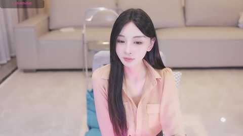 Media: Video of a young Asian woman with long black hair, fair skin, and a slender build, wearing a beige blouse, kneeling on a light blue cushion. Background features a beige couch and modern decor in a minimalist living room.