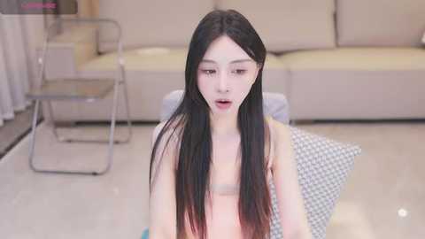 Media: Video of a young, slender East Asian woman with long black hair, fair skin, and small breasts, sitting on a checkered cushion in a minimalist, modern living room with beige furniture.