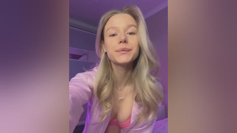 Media: Video of a young, fair-skinned, blonde woman with long hair, wearing a pink bra and unbuttoned shirt, smiling, in a dimly lit room with purple lighting.